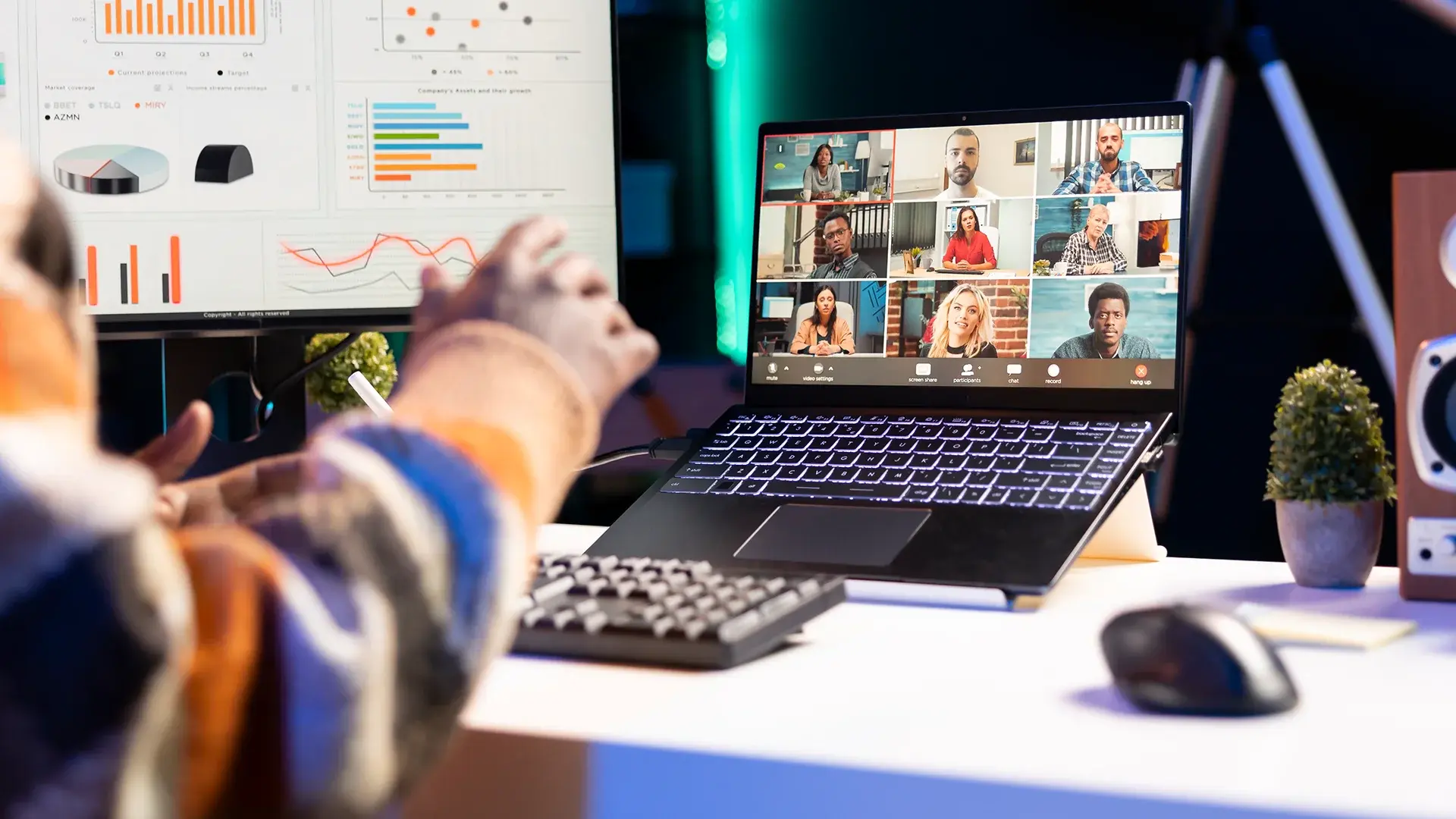 Person joining a multi-participant video webcast on a laptop at a desk, with business charts displayed on a monitor beside it.