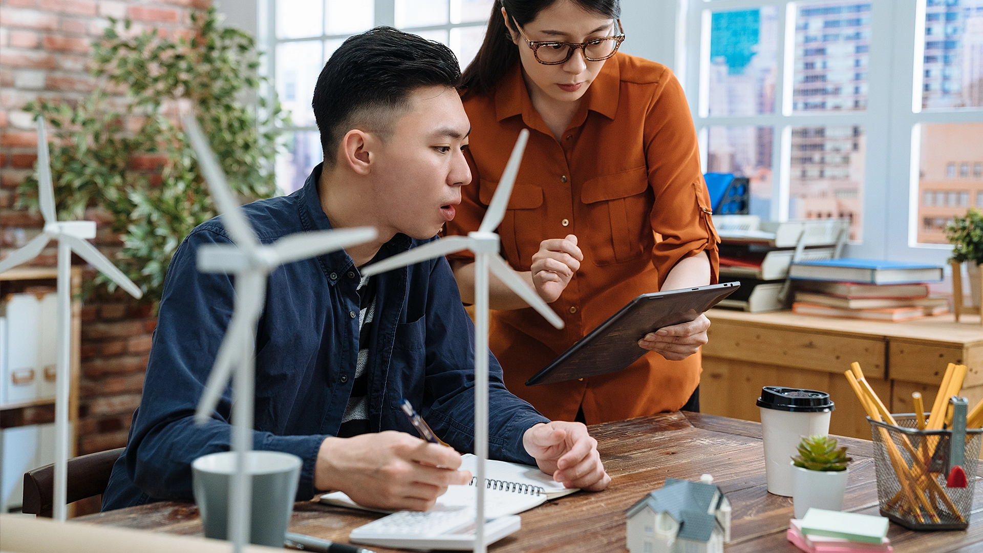 A man and woman collaborate in an office with wind turbine models, discussing energy topics on a tablet—ideal for an internal video knowledge base on energy solutions