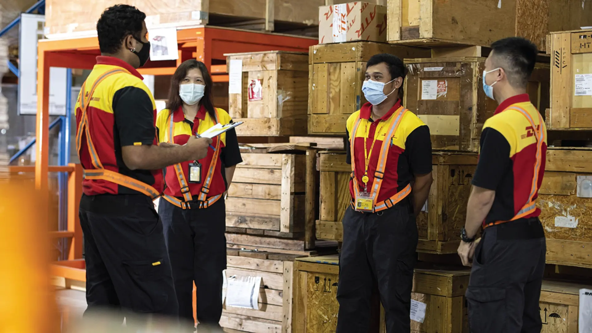 DHL India warehouse team in branded uniforms and safety gear conducting a team briefing, surrounded by wooden cargo crates, reflecting DHL’s commitment to logistics excellence and teamwork in India