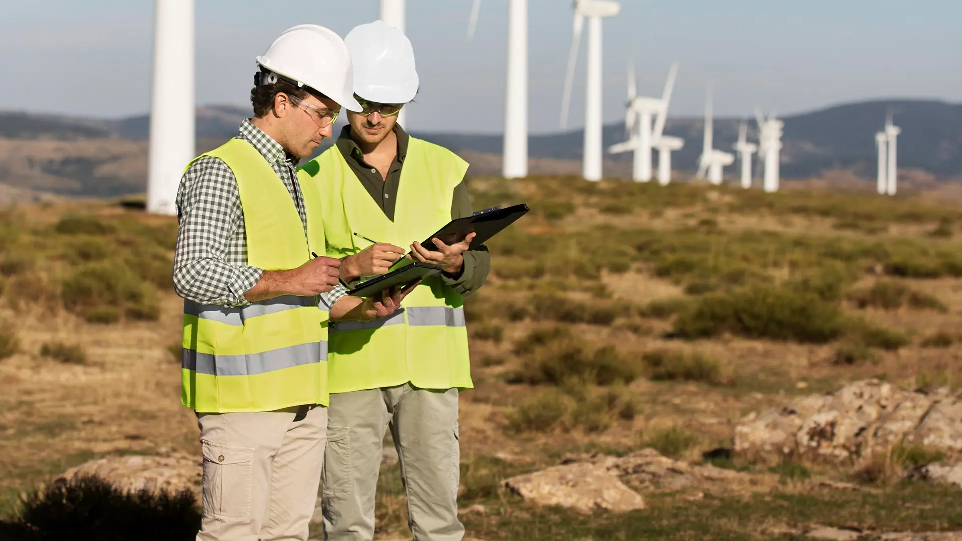 Two consultants wearing hard hats and safety vests reviewing documents in a wind farm, with multiple wind turbines in the background