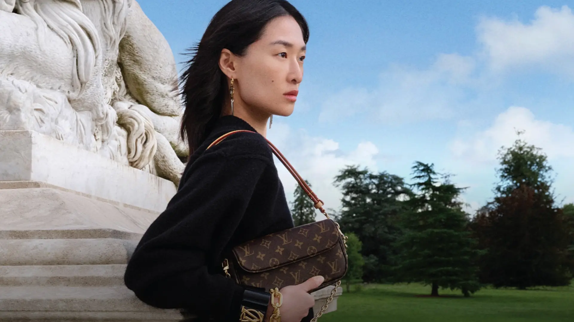 A woman stands outdoors, holding a Louis Vuitton monogrammed handbag, exuding elegance and sophistication against a scenic background