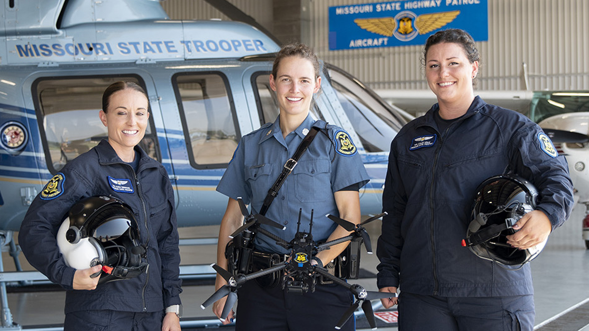 Three Missouri State Troopers, including two aviation officers holding helmets and a trooper with a drone, stand in front of a helicopter, symbolizing the Missouri Department of Public Safety’s law enforcement and aerial operations