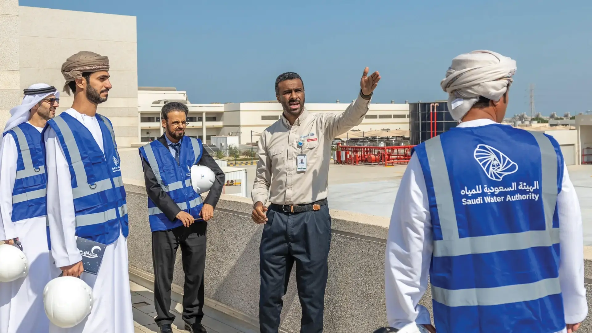 A group of officials from the Saudi Water Authority, wearing blue vests and helmets, engage in a discussion during an outdoor facility tour, representing the organization's commitment to water management and infrastructure