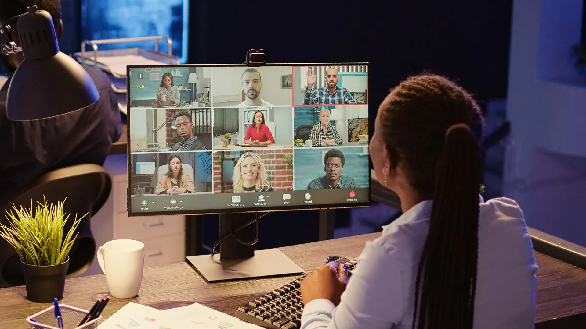 Employee on a video conference call with nine remote participants displayed in a grid on a desktop monitor.